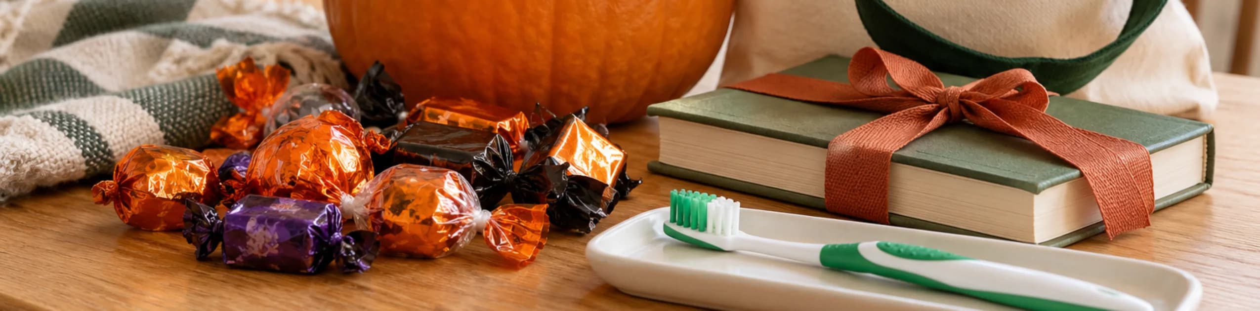 Pumpkin, wrapped Halloween candy, a toothbrush and a ribbon-tied book on a wooden table.